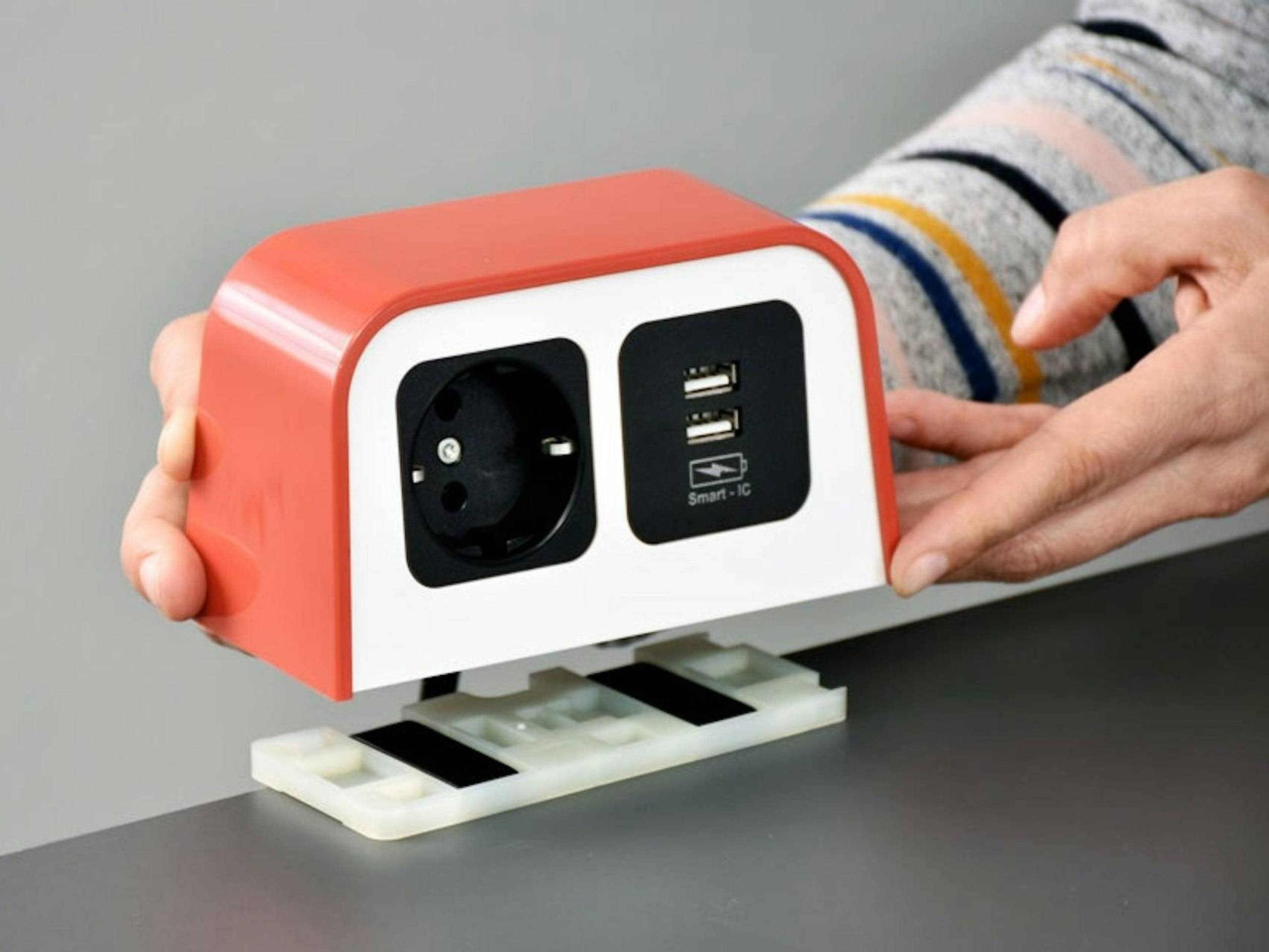 Rood-witte bureau-stopcontactdoos met één Europees stopcontact en twee USB-poorten, vastgezet op een bureau terwijl iemand het apparaat plaatst.
